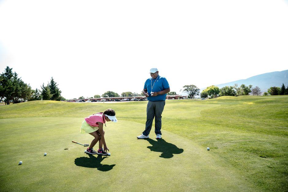 A young girl receives golf instruction on a sunny course from an expert, under clear skies.