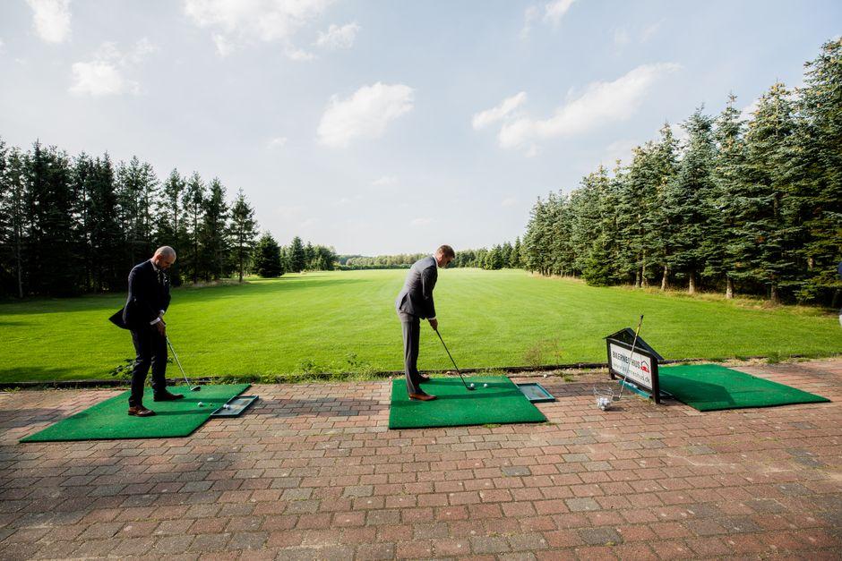Two men in suits playing golf on a lush, green course surrounded by trees on a sunny day.