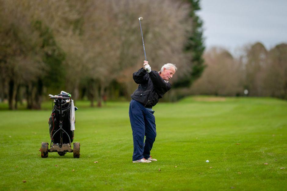 Elderly man swinging a golf club on a vibrant golf course with trolley in background.