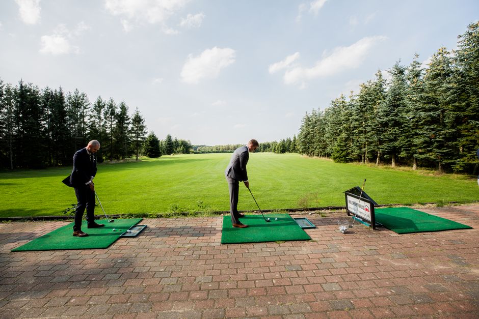 Two men in suits playing golf on a lush, green course surrounded by trees on a sunny day.